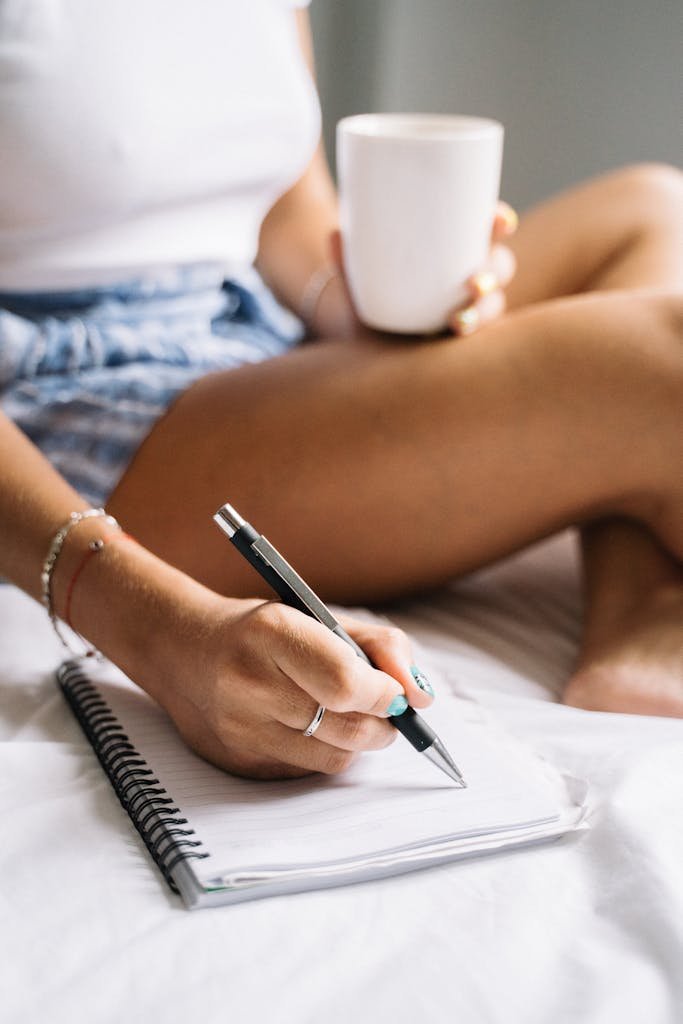 A woman enjoying a calm morning routine for success, drinking coffee and writing in a notebook while sitting in bed.