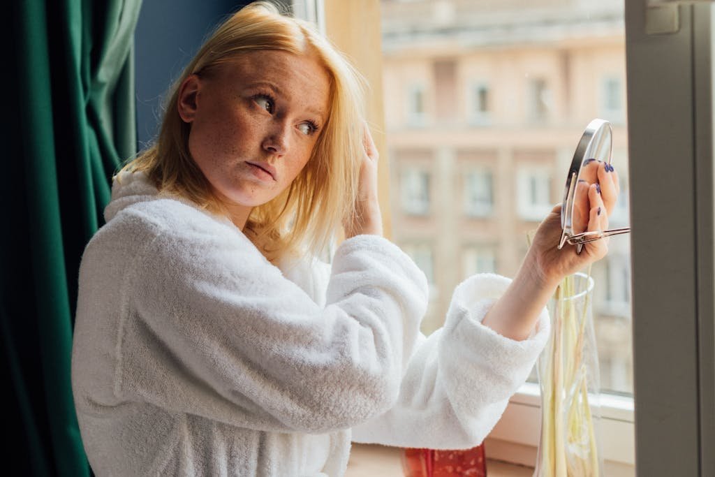 A woman in a white bathrobe looking in a mirror by the window.