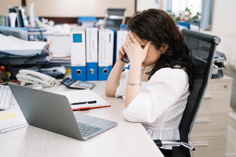 A woman in an office sitting at a desk, visibly stressed, using a phone. Workplace stress concept.
