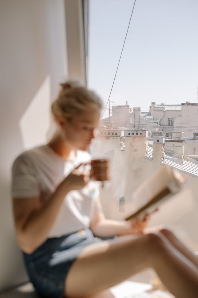 A woman sits and reads by a window, with a view of rooftops and clear skies.