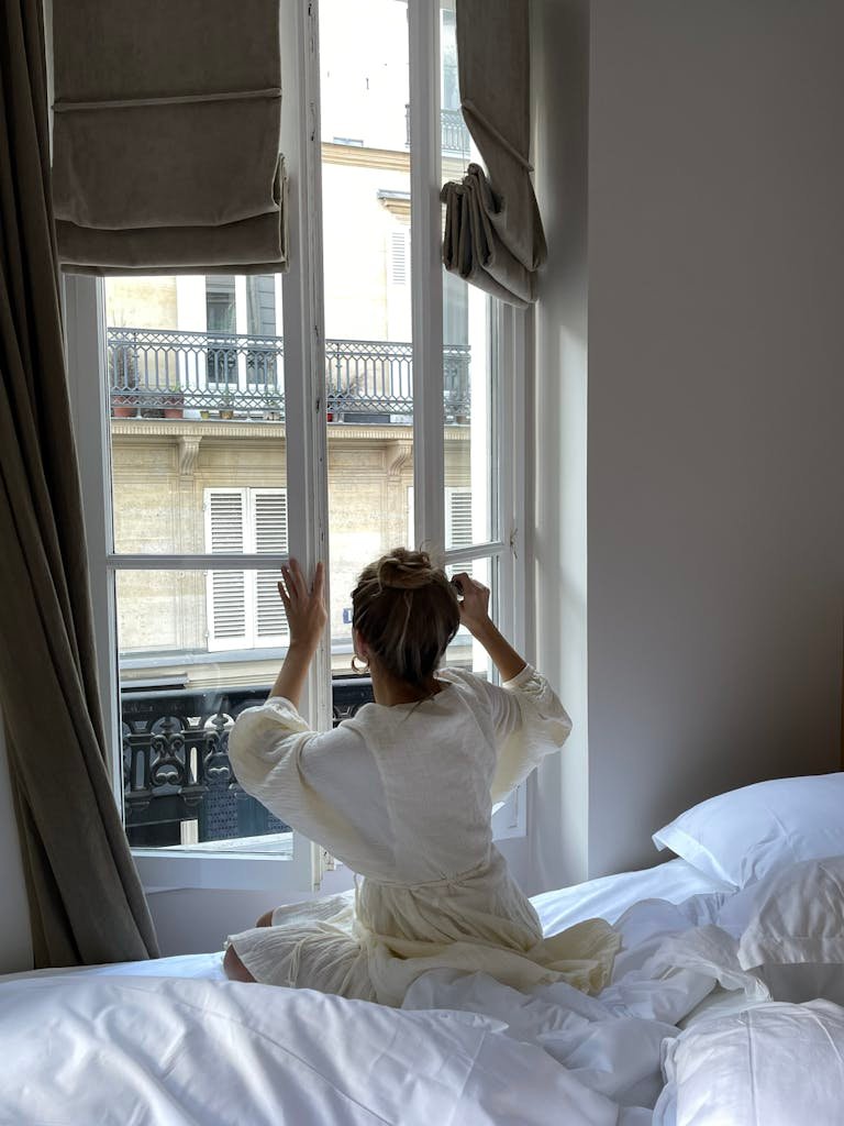 A woman sitting on a bed opens a window in a Parisian bedroom with natural light streaming in.