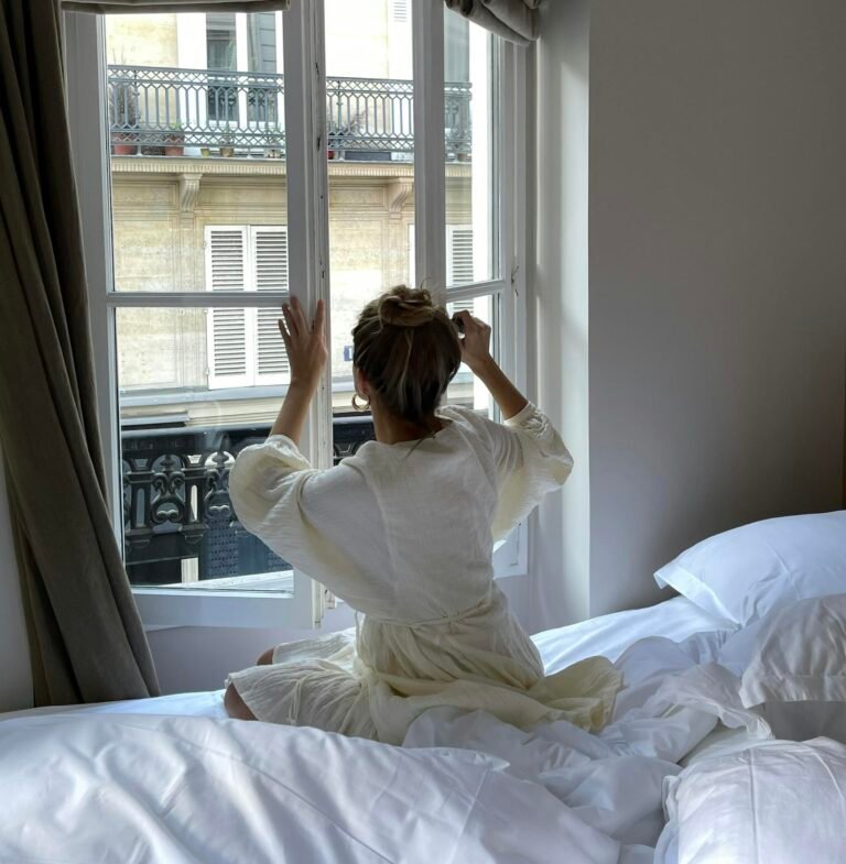 A woman sitting on a bed opens a window in a Parisian bedroom with natural light streaming in.