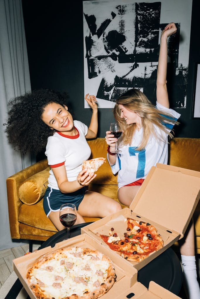 Two friends enjoying a fun pizza party indoors, sharing laughter and red wine.