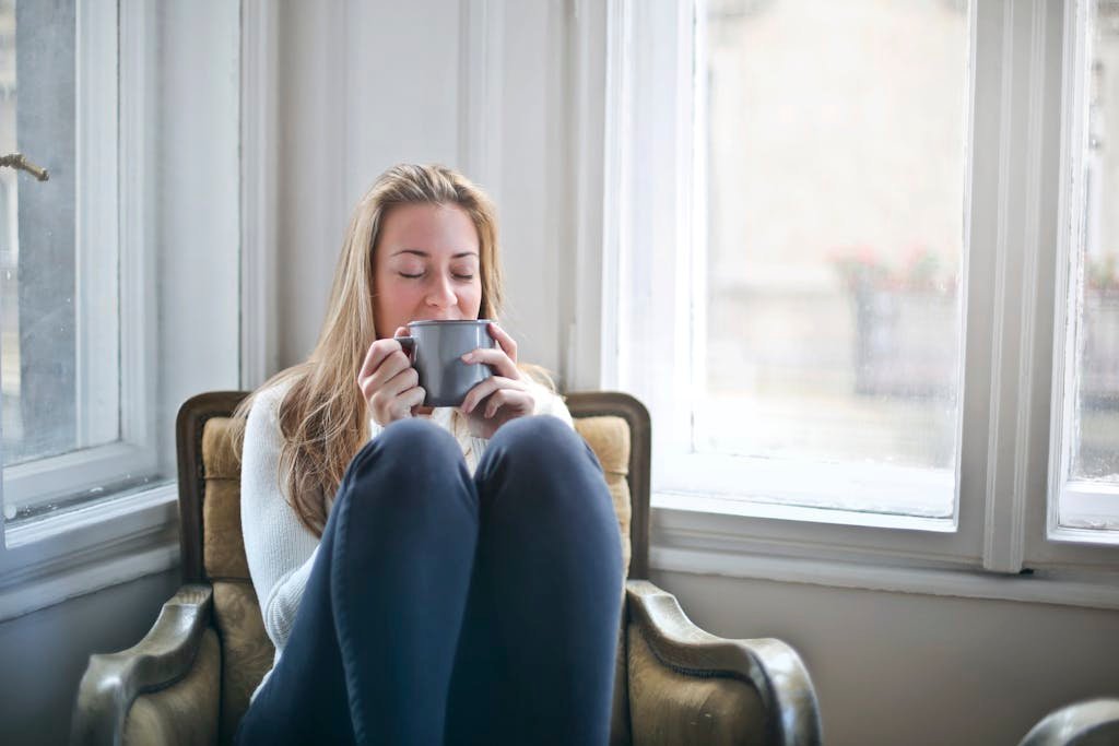 Woman enjoying a warm drink in a cozy armchair by a window, embracing relaxation.
