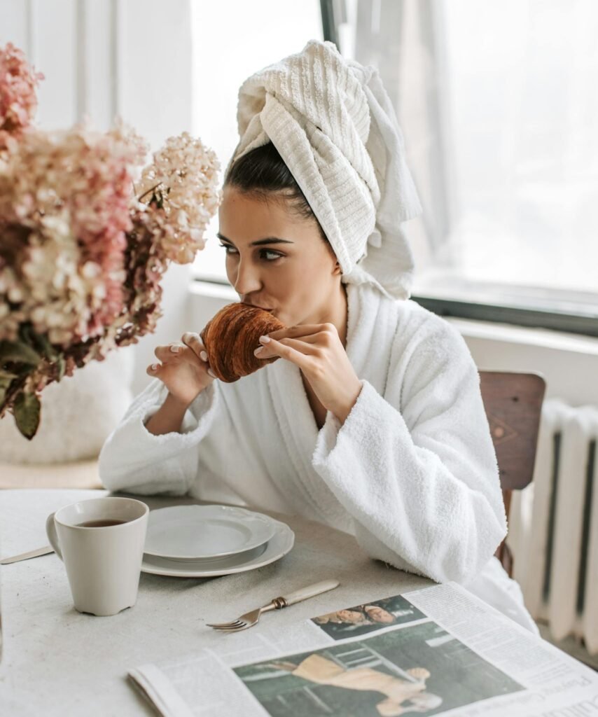 Woman in bathrobe eats croissant with coffee at sunny dining table.