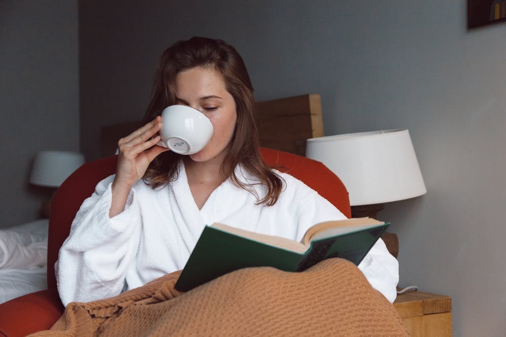 Woman in bathrobe enjoying coffee and reading a book in a cozy bedroom setting, capturing a peaceful moment.