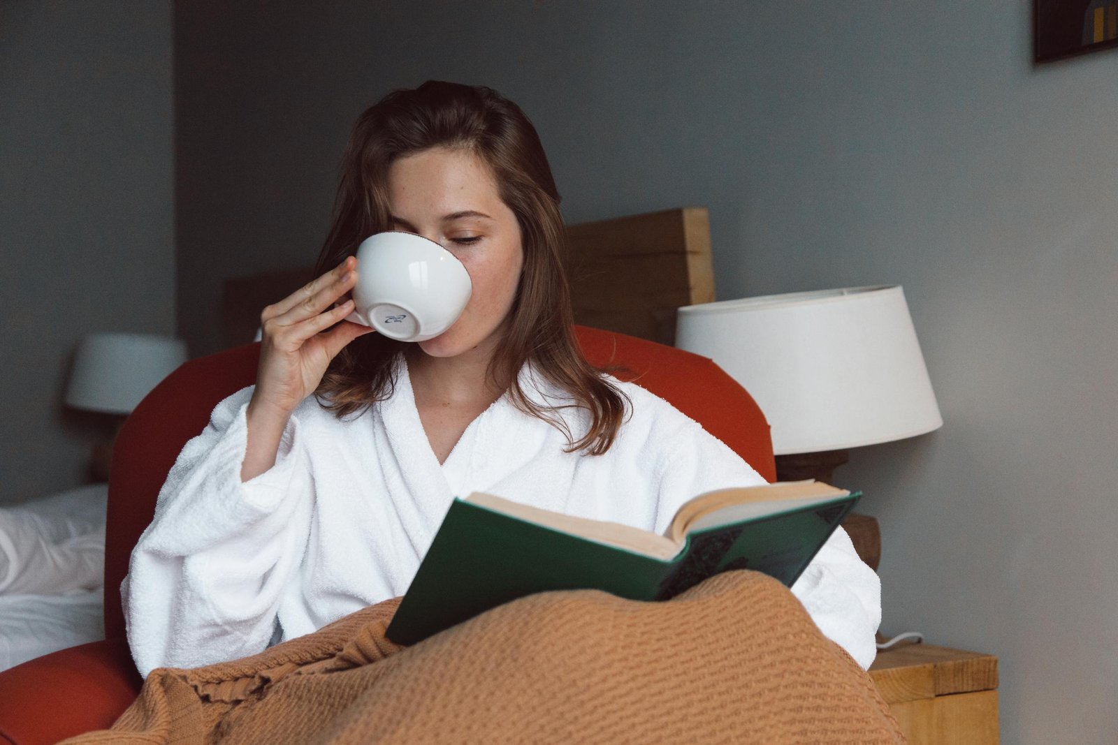 Woman in bathrobe enjoying coffee and reading a book in a cozy bedroom setting, capturing a peaceful moment.