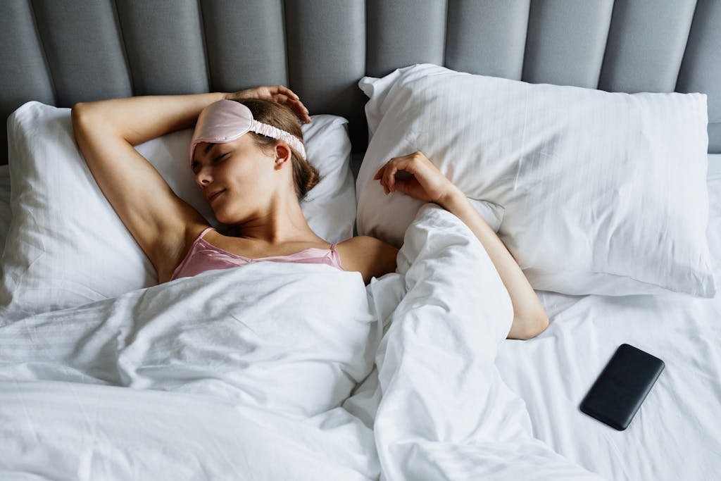 Woman resting in bed wearing an eye mask with a smartphone nearby, embracing relaxation.