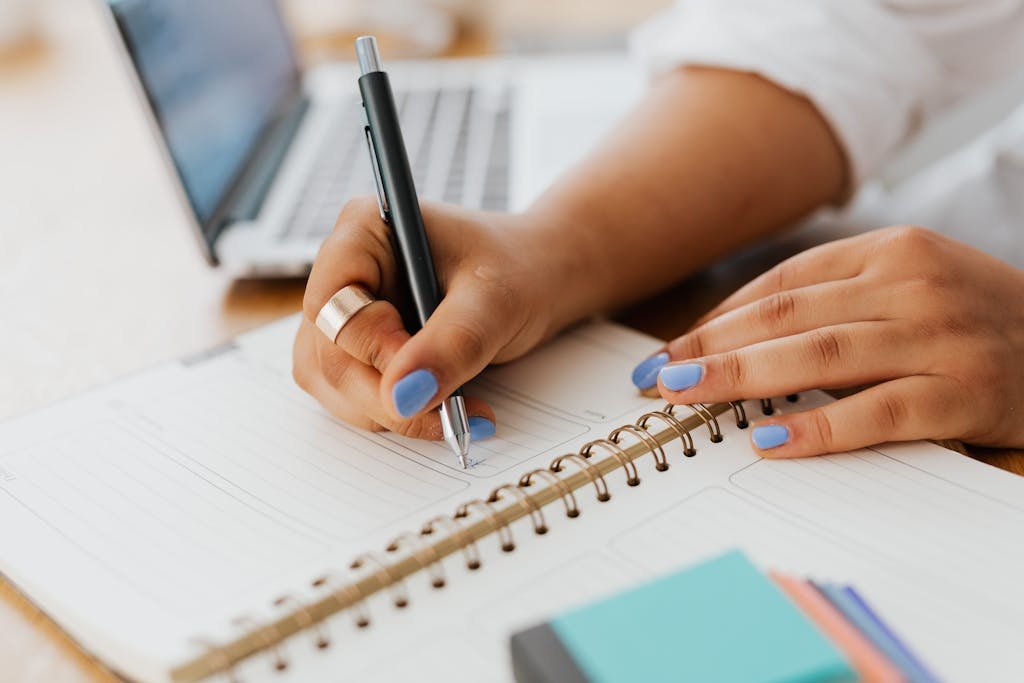 Woman with blue nails writing in a spiral notebook beside a laptop, depicting a work or study setting.