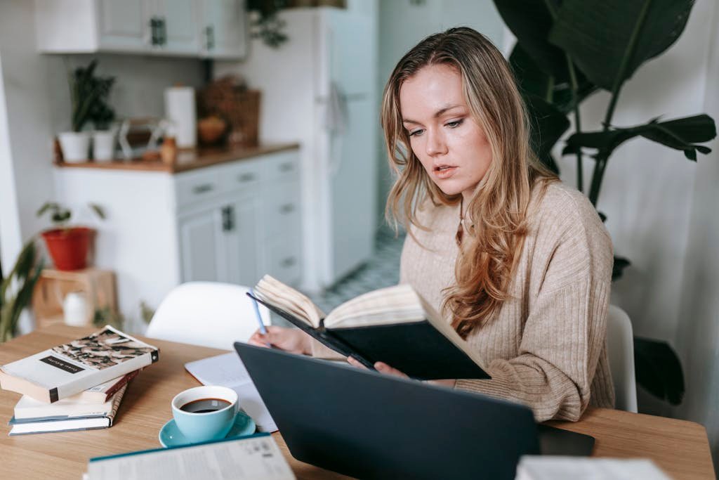 Young focused female studying planner near netbook and cup of hot aromatic coffee on blurred background of kitchen interior