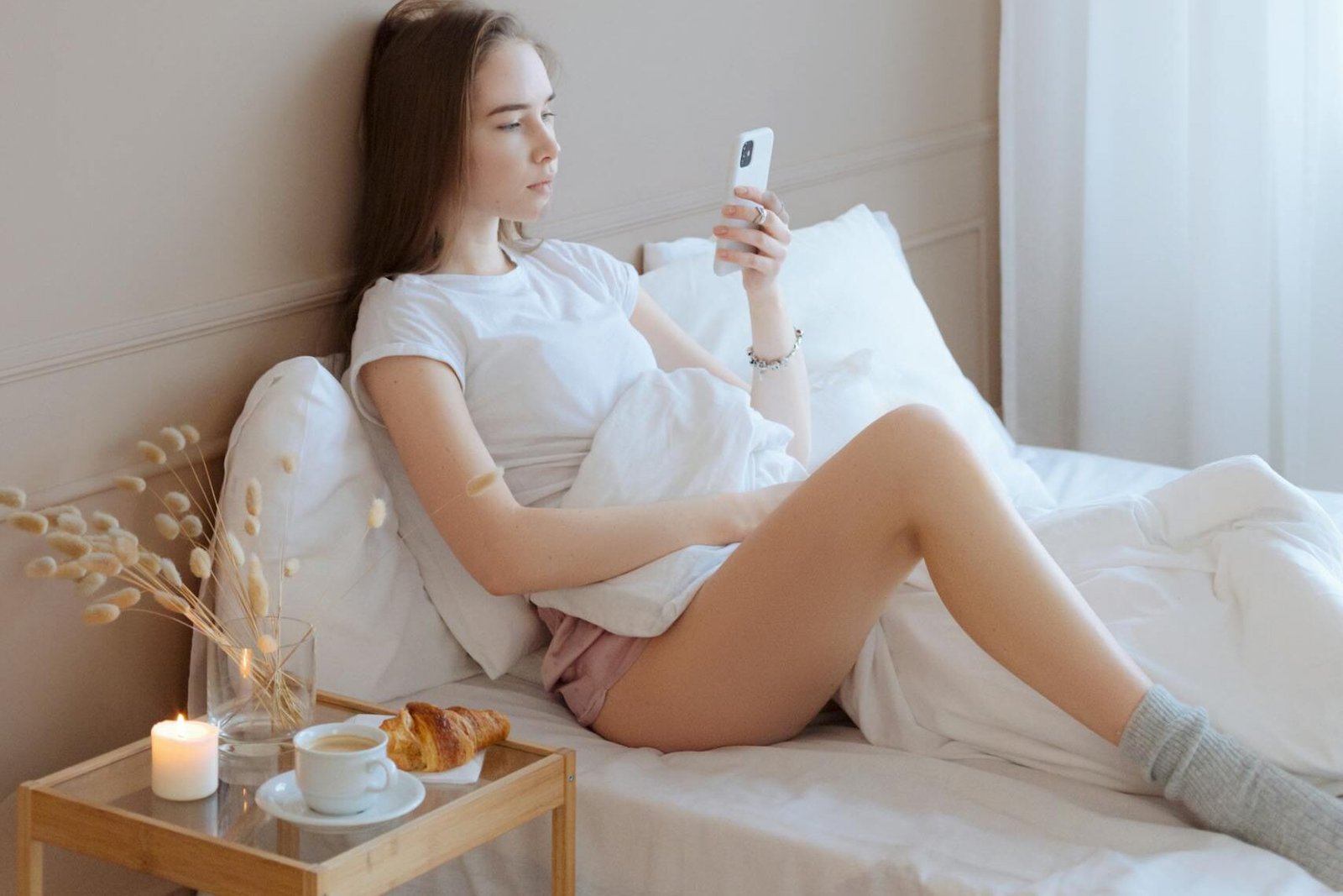 Young woman enjoys breakfast in bed, sipping coffee and using her phone in a peaceful morning setting.