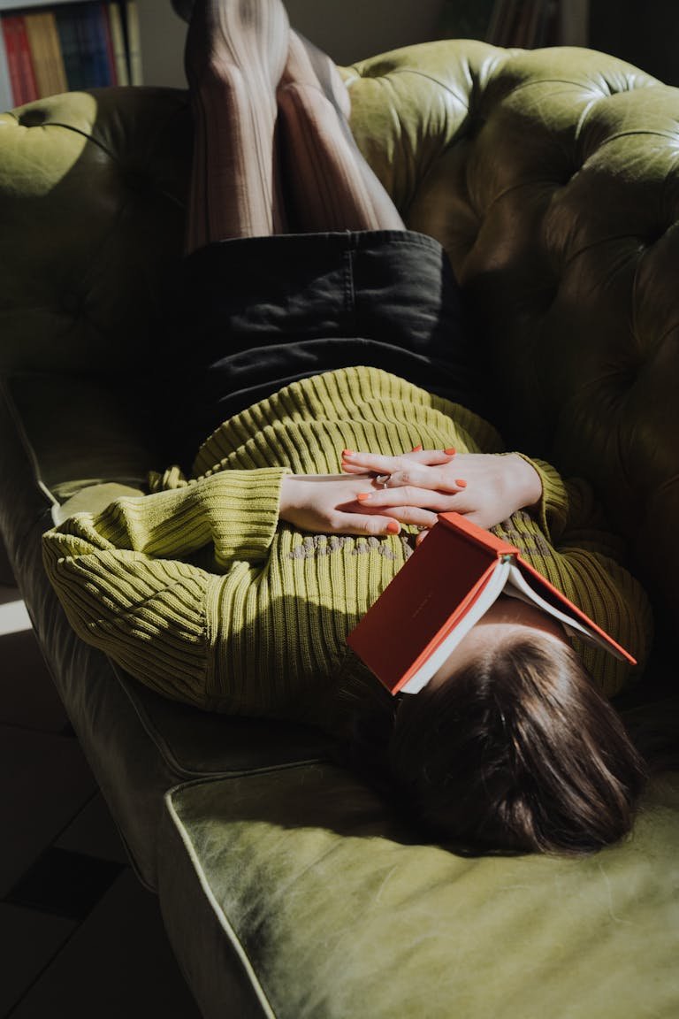 Young woman in green sweater and mini skirt relaxing with book on couch indoors.