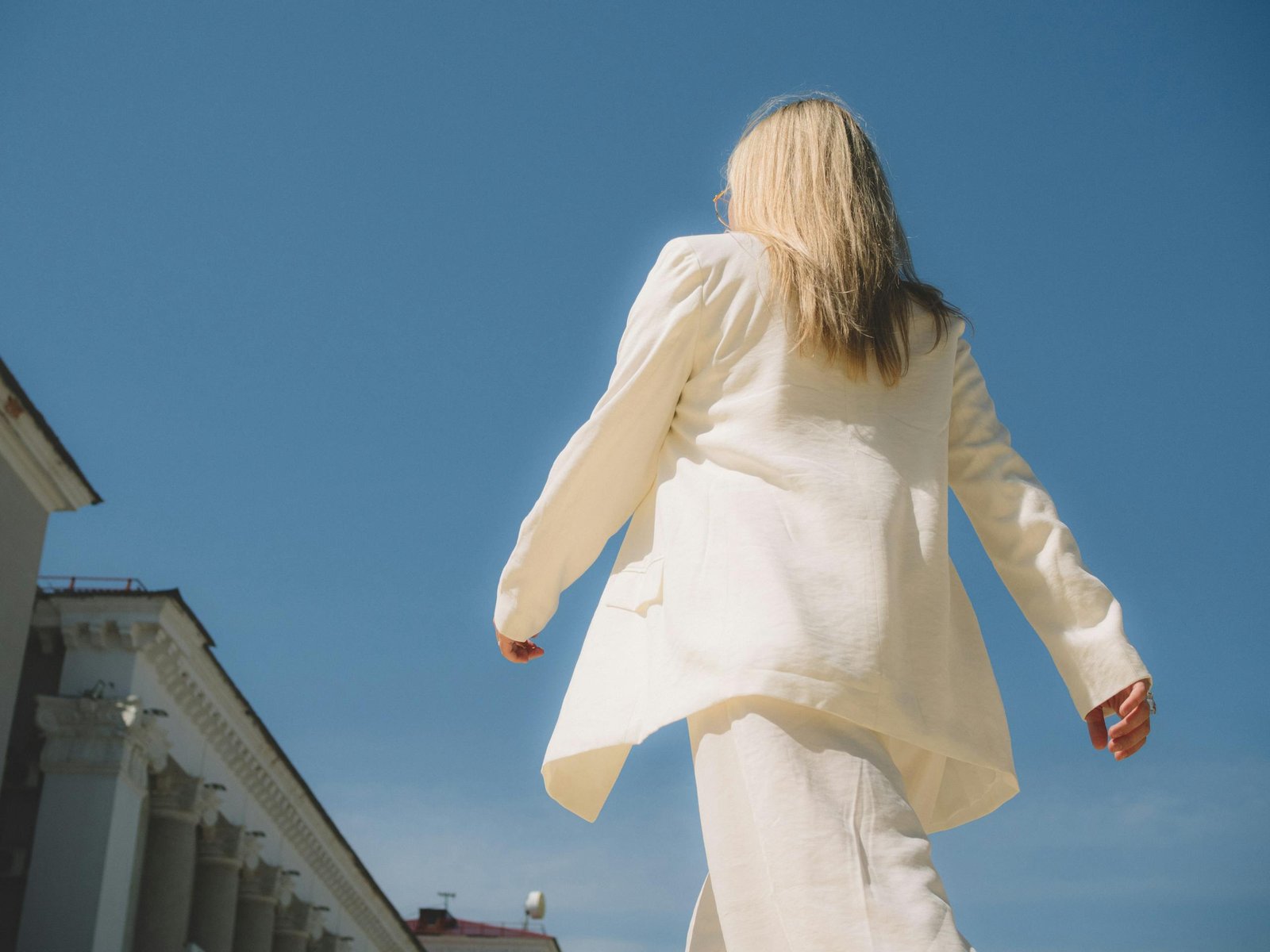 A woman in white formal wear walking outside with an architectural backdrop.