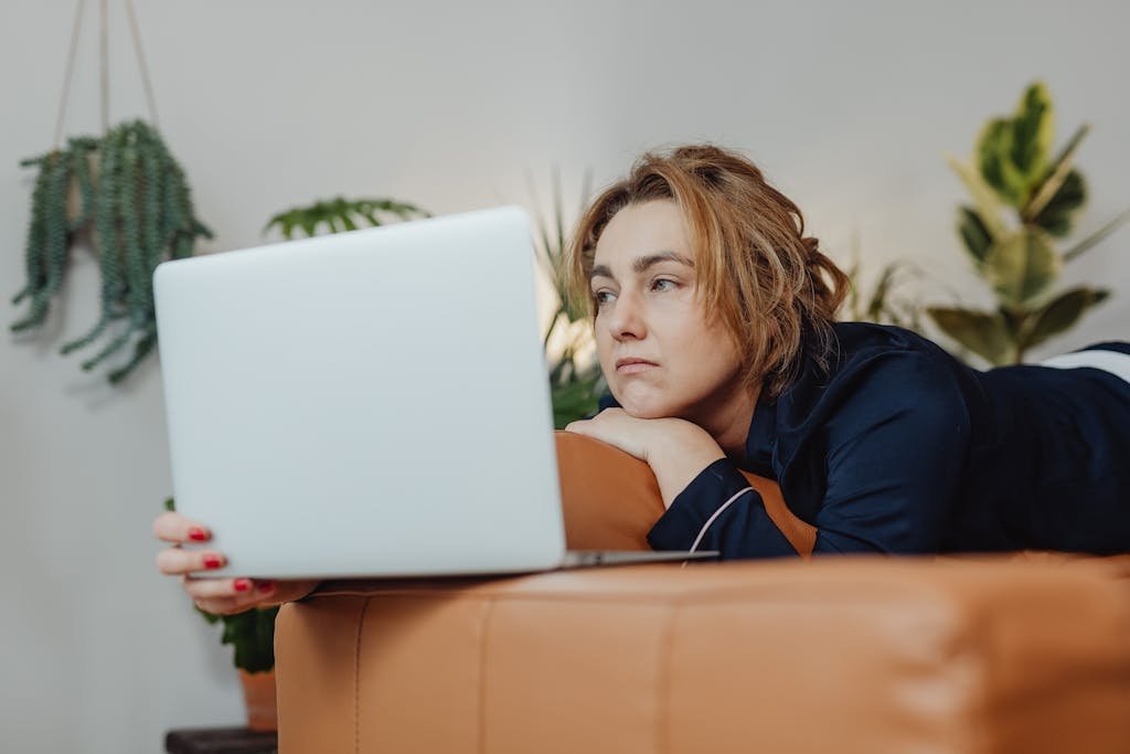 A woman lying on a couch indoors, looking upset while holding a laptop in a cozy home environment.