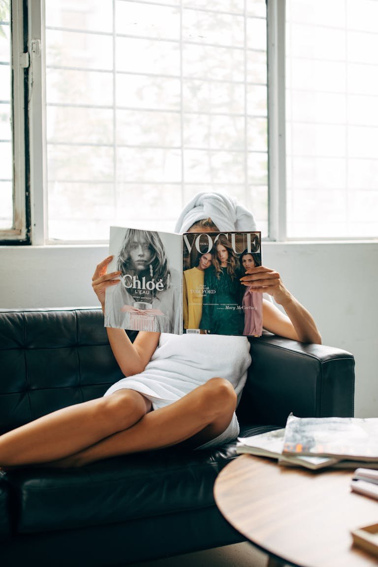 A woman with a towel wrapped on her head reads a Vogue magazine while lounging on a couch.