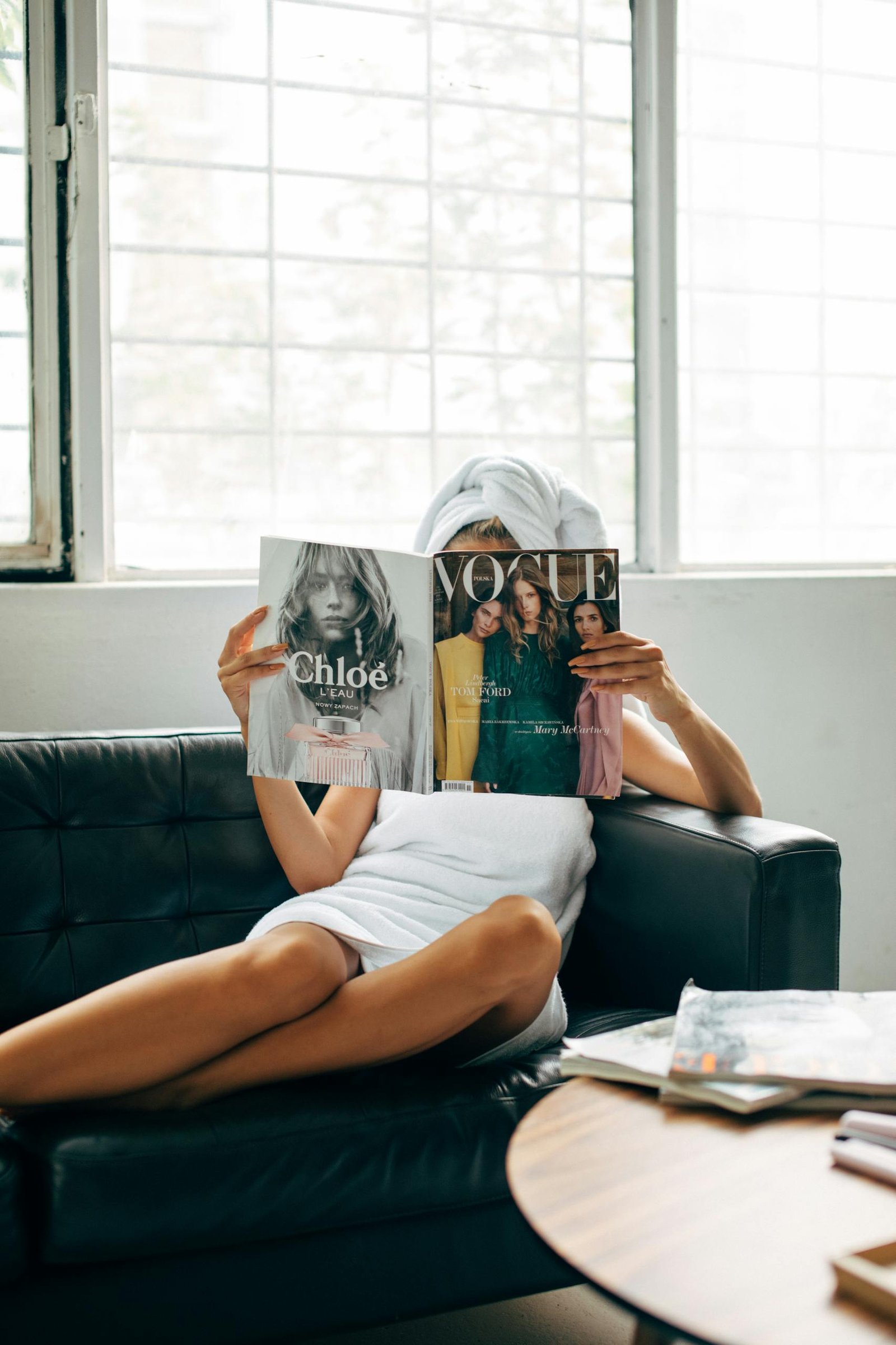 A woman with a towel wrapped on her head reads a Vogue magazine while lounging on a couch.