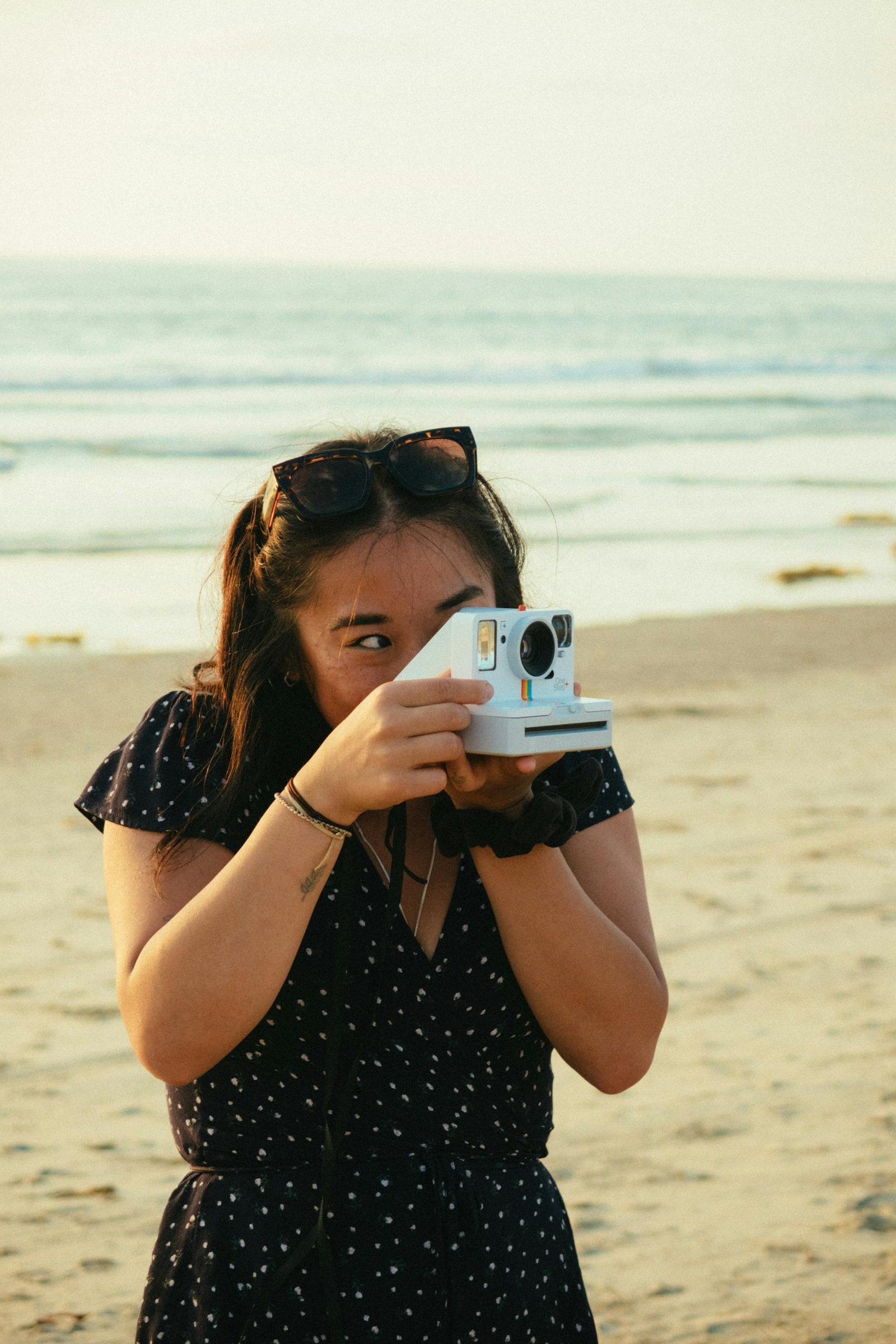 A woman with sunglasses practicing her hobby as she takes a picture on a beach using a Polaroid camera.