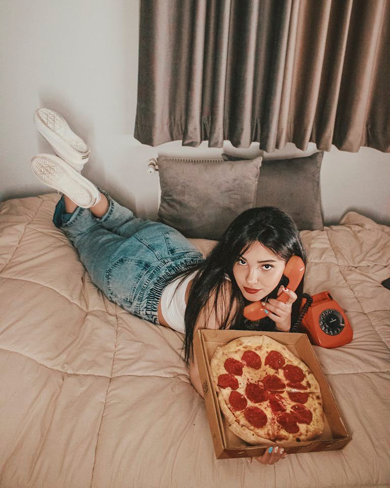 A young woman casually lying on a bed talking on a retro phone while enjoying a pizza.