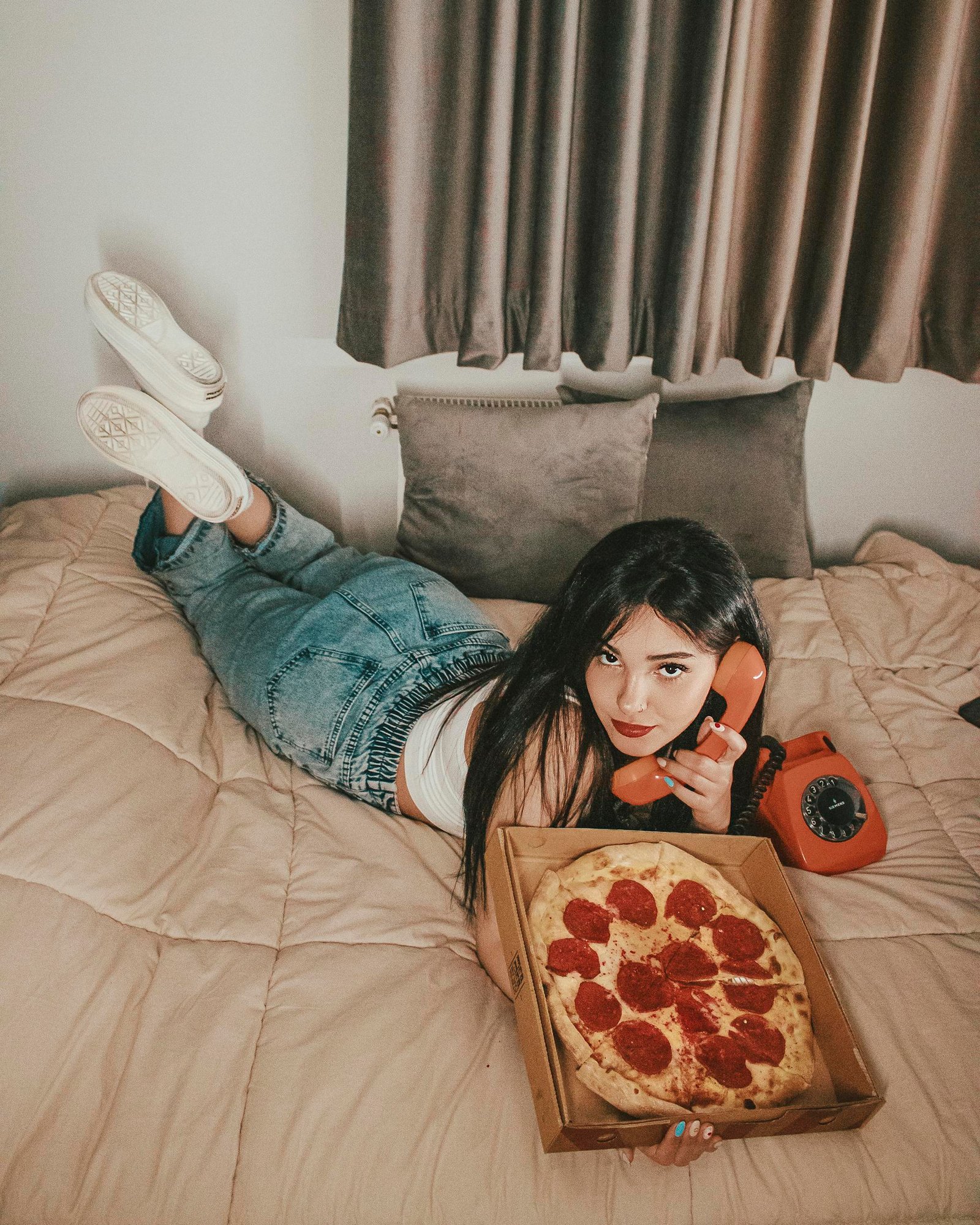 A young woman casually lying on a bed talking on a retro phone while enjoying a pizza.
