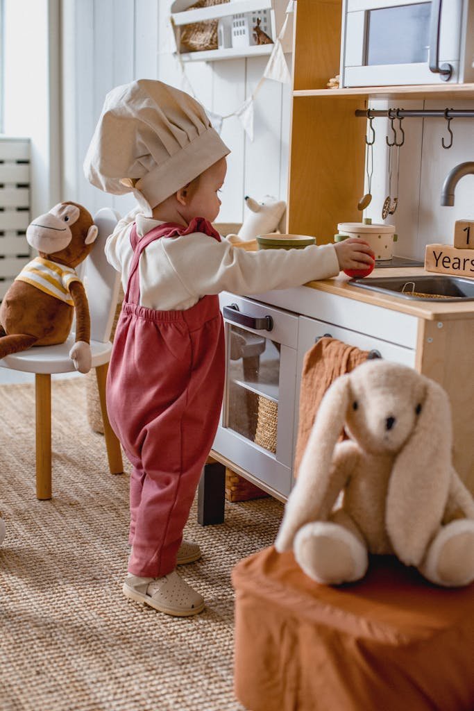 Adorable child wearing a chef hat playing in a toy kitchen with stuffed animals.