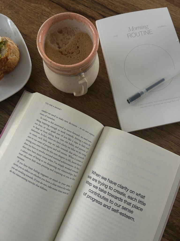 Morning routine journaling setup with coffee, an open book, and a minimalist daily journal on a wooden table.