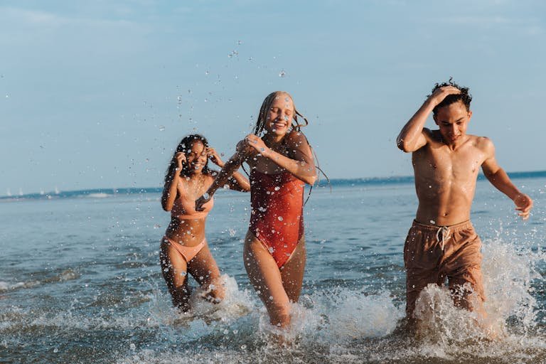 Three teenagers enjoy a playful day at the seaside, splashing through the waves under a clear summer sky.
