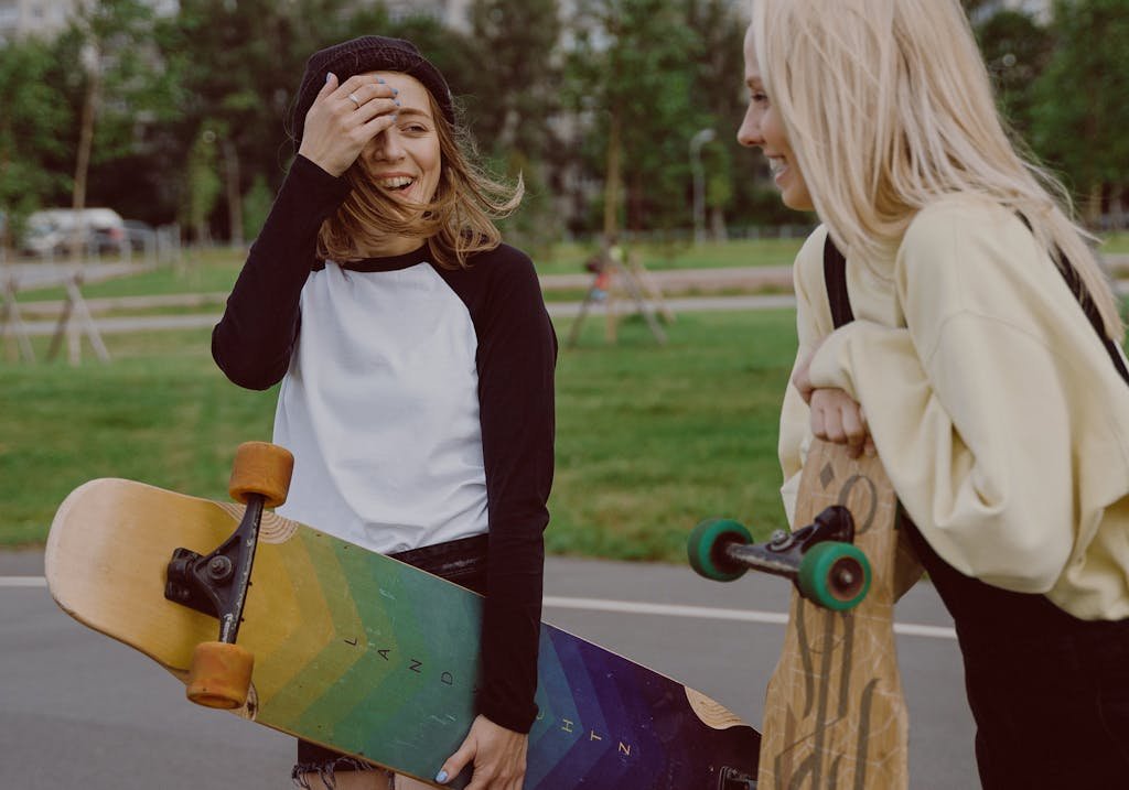 Two teenage girls laughing and holding longboards in a park, enjoying leisure time together.