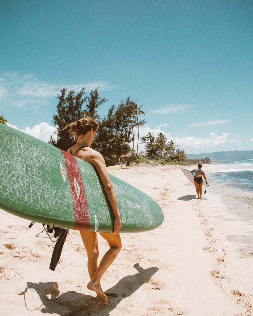 Two women walking on a sandy Hawaiian beach carrying surfboards under a clear blue sky.