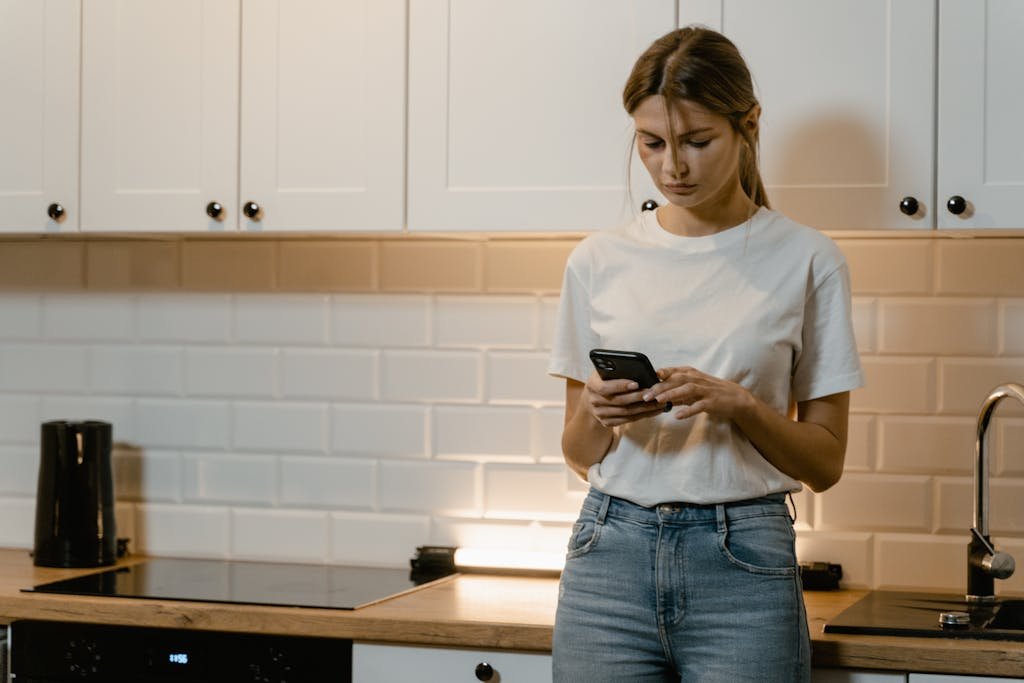 Young woman in kitchen using smartphone, looking focused and thoughtful.