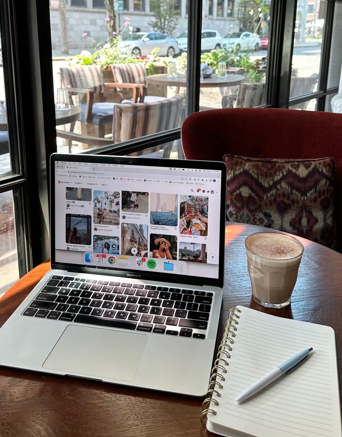 Laptop open on a café table beside a notebook and a warm drink, creating a cozy setting for working on a side hustle with natural light coming through large windows.