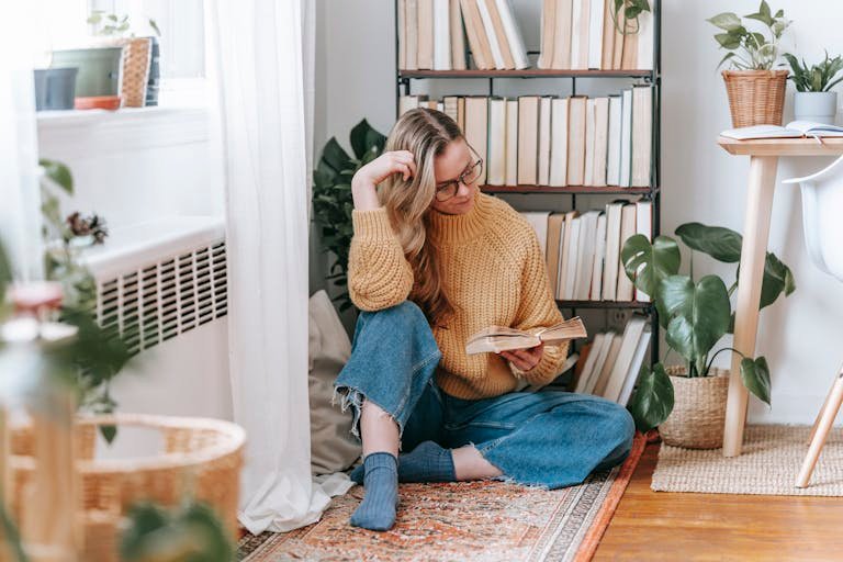 Woman reading in a cozy corner with plants and books, wearing a knitted sweater.