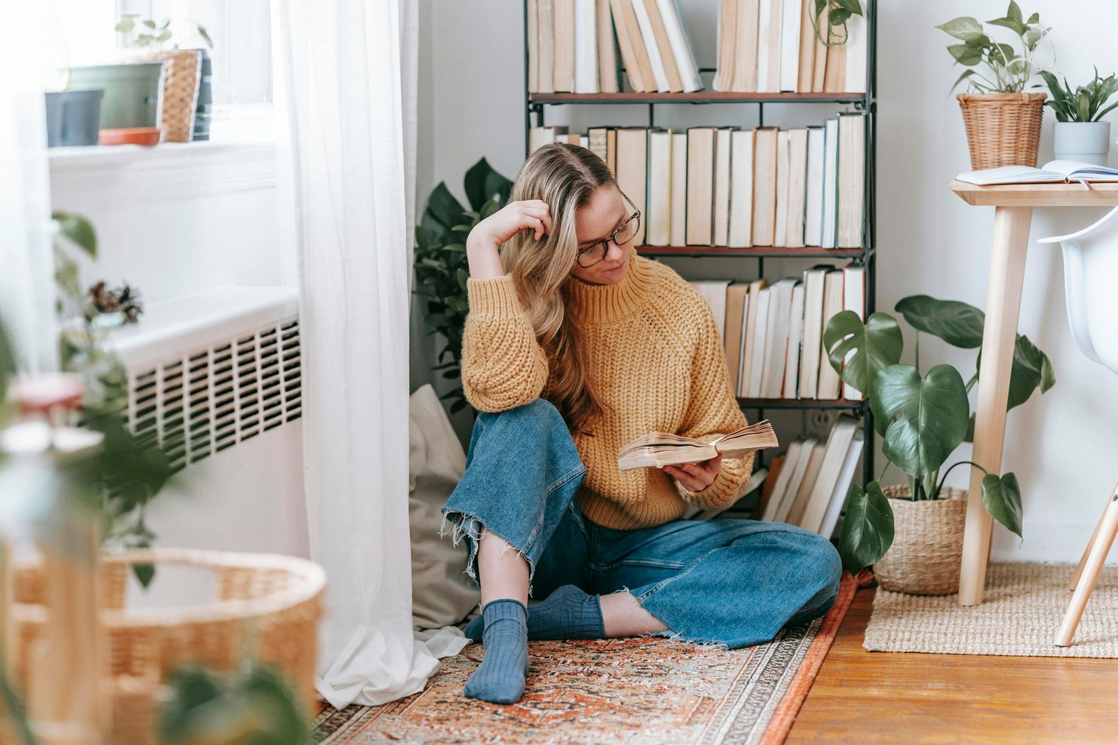 Woman reading in a cozy corner with plants and books, wearing a knitted sweater.
