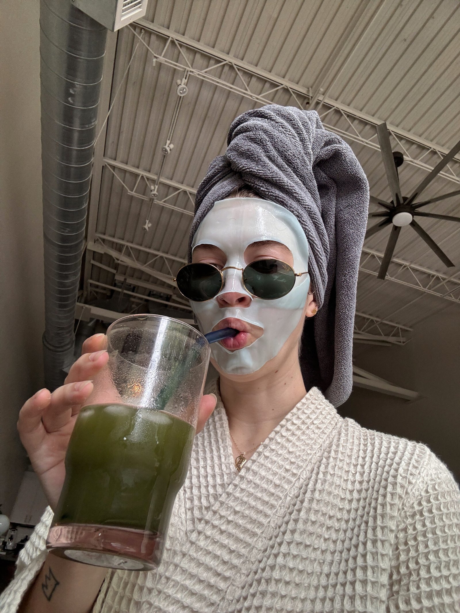 Woman in a bathrobe with a towel on her head, wearing a face mask and drinking green juice during a relaxed, that girl morning routine.