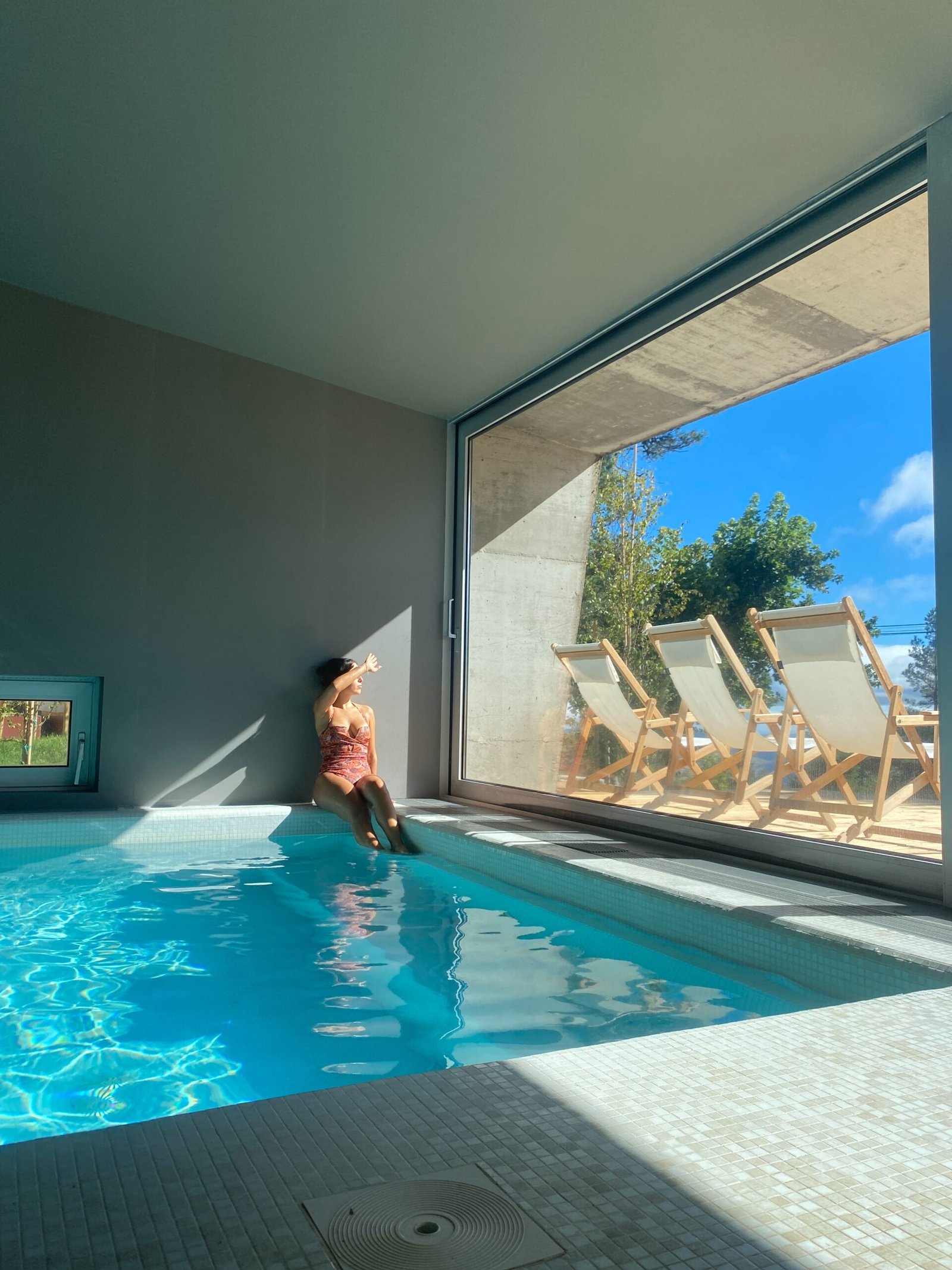 Woman sitting by an indoor pool with sunlight streaming through a large window, creating a calm, cozy summer atmosphere indoors.